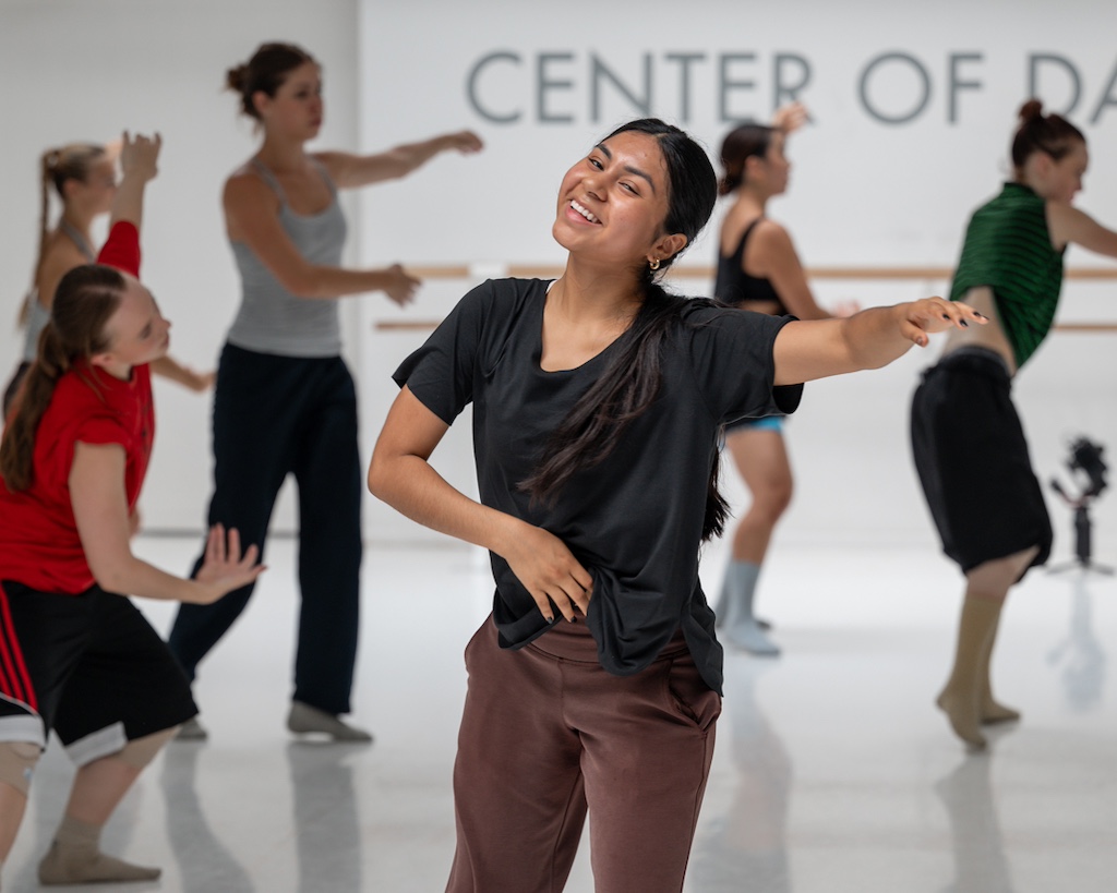 A dancer smiles in mid-practice with other dancers behind her during a training session in a dance studio